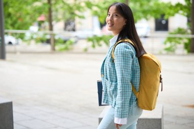Chinese student wearing a blue jacket and yellow backpack is walking down a sidewalk. She is smiling and she is in a good mood