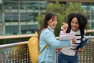 Two young women are standing on a bridge, smiling and holding books. They seem to be enjoying each others company and discussing their studies