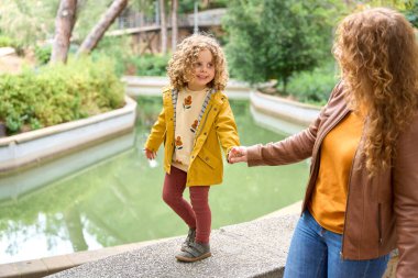 Bir kadın, bir su kütlesinin yanından geçerken bir çocuğun elini tutuyor. Çocuk sarı bir ceket ve çiçekli bir gömlek giyiyor. Gökyüzünü yansıtan suyla sahne huzurlu ve huzurlu.