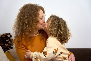 Bir anne ve kızı birbirlerine sarılıyorlar. Anne turuncu bir kazak giyiyor ve kız çiçekli bir gömlek giyiyor.