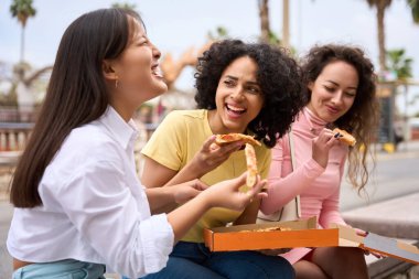 Üç kadın bir bankta oturmuş pizza yiyorlar. Gülüyorlar ve yemeklerini yiyorlar.