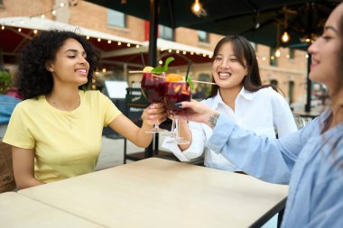 Üç kadın bir masada oturuyor, her biri bir bardak şarap tutuyor. Gülüyorlar ve birbirlerine kadeh kaldırıyorlar.