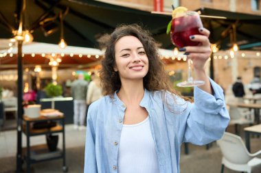 Bir kadın bir bardak şarap tutuyor ve gülümsüyor. O, dışarıda bir restoranda duruyor. Atmosfer rahat ve rahat.