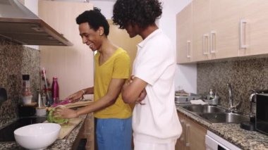 İki adam mutfakta birlikte yemek pişiriyor. İçlerinden biri sarı bir gömlek giyiyor. Salata hazırlıyorlar.