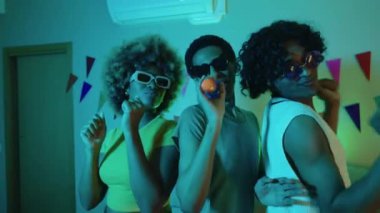 LGBTQIA arkadaşları bir ev partisinde renkli ışıklar altında dans edip şarkı söylüyor.