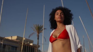 Kavis, kumlu bir sahilde palmiye ağaçları ve teknelerle çevrili kırmızı bikinili kıvırcık saçlı bir kadını yakalıyor.