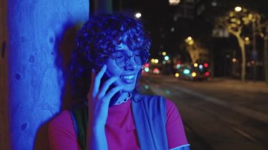 Genç, kızıl saçlı, renkli gözlüklü bir kadın telefonda konuşurken gece vakti şehirde ona parlak renkler saçan görünmez neon ışıkların altında dikiliyor.