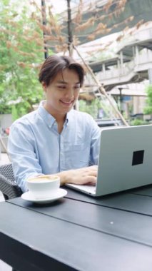 Young freelancer is smiling while typing on a laptop and drinking coffee at a table in bangkok