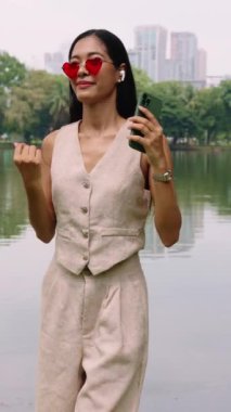 Kalp şeklinde güneş gözlüğü takan nüfuzlu biri, Bangkok parkında telefonuna yavaş çekimde poz veriyor.