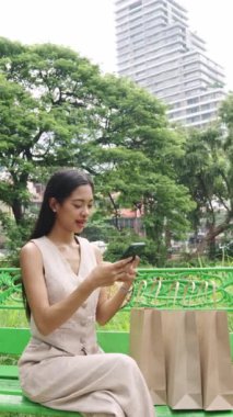 Genç bir kadın akıllı telefonunu Bangkok 'ta bir bankta yanında alışveriş torbalarıyla ağır çekimde kullanıyor.