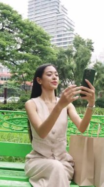 Genç bir kadın bankta oturuyor ve akıllı telefonunu ağır çekimde kullanıyor.