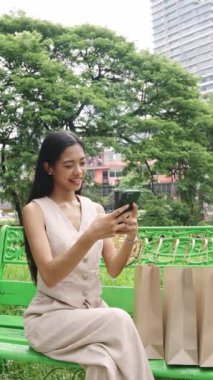 Genç bir kadın, yanında alışveriş torbalarıyla Bangkok parkında telefonunu yavaş çekimde kontrol ediyor.