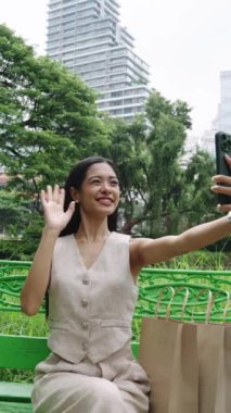 Bej renkli yelek giyen genç bir kadın bankta oturuyor yanında alışveriş torbalarıyla selfie videosu çekiyor.