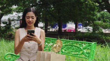 Genç bir kadın parkta bir bankta oturuyor, yanında alışveriş torbalarıyla akıllı telefonunu kullanıyor.