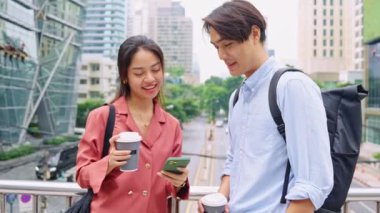 İki genç profesyonel, bir şehir köprüsünde kahve molası verip sohbet ediyorlar ve akıllı telefonu yavaş çekimde kullanıyorlar.