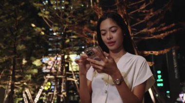 Asyalı iş kadını elinde akıllı bir telefon tutuyor, gece kalabalık bir bangkok caddesinde uygulamaları ve sosyal medyayı tarıyor.