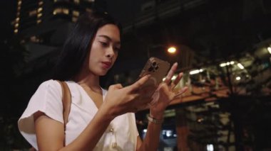 Genç iş kadını geceleri Bangkok caddesinde akıllı telefonunu kullanıyor ve canlı şehir atmosferinin tadını çıkarıyor.