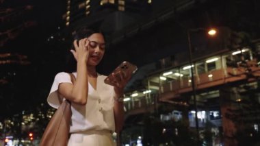 Genç iş kadını gece Bangkok 'ta bir sokakta dikiliyor, akıllı telefonunu kullanırken gülümsüyor.