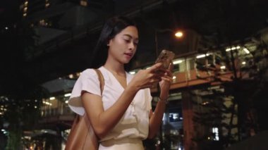 Genç iş kadını gece bir bangkok caddesinde duruyor, şehir ışıklarının arasında akıllı telefonuna odaklanmış.