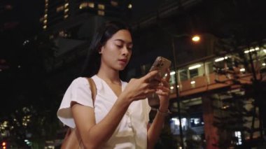 Genç iş kadını gece canlı Bangkok 'ta akıllı telefonunu karıştırırken gülümsüyor, şehir atmosferinin tadını çıkarıyor.