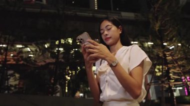 Genç iş kadını geceleri Bangkok sokaklarında telefonuyla gülümsüyor.