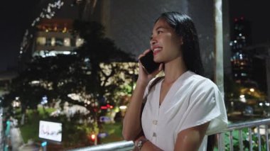 İş kadını gece Bangkok 'ta bir köprüde duruyor, telefonuyla konuşuyor ve kendine gülerek gülümsüyor.