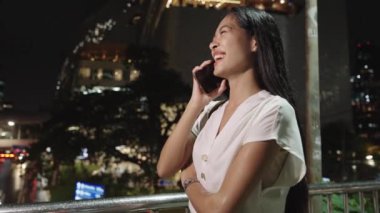 İş kadını, Bangkok 'ta gece vakti bir köprüde konuşurken gülüyor. Şehir ışıklarıyla çevrili.