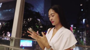 Geceleri Bangkok 'ta akıllı telefonunu kullanarak gülümseyen genç iş kadını, canlı şehir atmosferinin ve teknolojisinin tadını çıkarıyor.