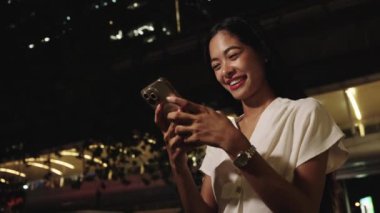 Gece canlı bir Bangkok şehrinde duran iş kadını, akıllı telefonuna bakarken gülümsüyor.