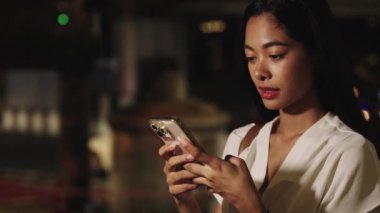 Genç iş kadını gece Bangkok 'un canlı sokaklarında akıllı telefonunu kullanırken gülümsüyor, şehir ışıklarıyla çevrili.