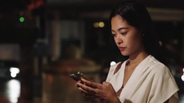 Genç iş kadını gece Bangkok 'un canlı sokaklarında akıllı telefonunu kullanırken gülümsüyor.