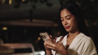 Genç iş kadını gece Bangkok 'ta kaldırımda dikiliyor, akıllı telefonuna bakarken gülümsüyor.