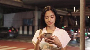 İş kadını gece bir sokak köşesinde duruyor, gülümsüyor ve Bangkok 'un canlı şehrinde akıllı telefonunu kullanıyor.