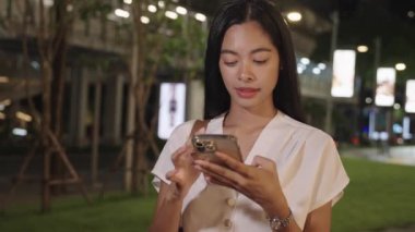 Genç iş kadını gece Bangkok 'ta kaldırımda dikiliyor, akıllı telefonuna bakarken gülümsüyor.