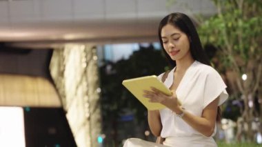 Gece Bangkok 'ta bir binanın önünde tablet üzerinde çalışan genç bir iş kadını. Etrafı şehir ışıkları ve ambiyansla çevrili.