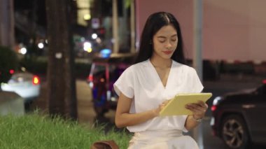 Bangkok 'ta bir bankta oturan genç iş kadını tablet kullanıyor ve şehrin atmosferinin tadını çıkarırken gülümsüyor.