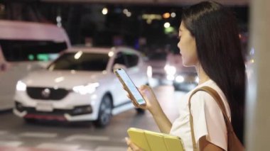 Genç Asyalı iş kadını Bangkok 'ta işlek bir caddenin kenarında beklerken telefonunda arama uygulaması kullanıyor.