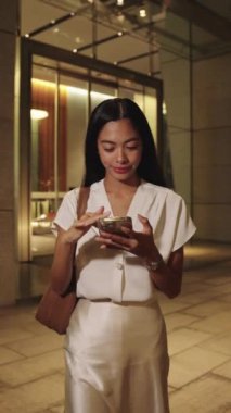 Genç iş kadını kaldırımda duruyor, Bangkok 'un canlı sokaklarında akıllı telefonunu karıştırıyor.