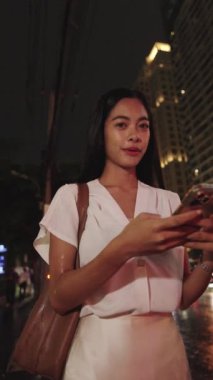 Genç iş kadını gece bangkok kaldırımında duruyor, akıllı telefonunu kullanıyor ve hareketli şehir atmosferinin tadını çıkarıyor.