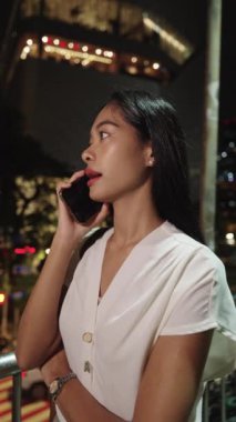 Genç iş kadını gece Bangkok 'ta bir köprüde duruyor, telefonla konuşuyor ve gülümsüyor.