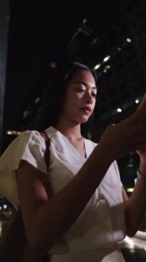 Bangkok 'ta gece akıllı telefon kullanırken gülümseyen iş kadını. Etrafı canlı şehir ışıkları ve modern kentsel ortam ile çevrili.