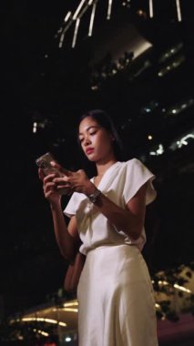 Genç bir iş kadını gece canlı bir Bangkok şehrinde duruyor, akıllı telefonuyla gülümsüyor.