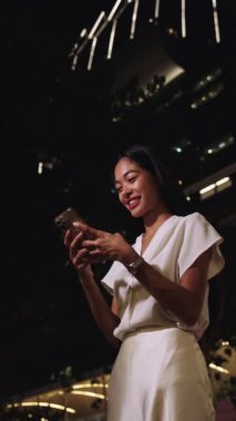 Bangkok 'ta gece akıllı telefon kullanan iş kadını gülümsüyor. Etrafı canlı şehir ışıkları ve modern gökdelenlerle çevrili.