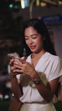 Gülümseyen genç iş kadını Bangkok 'ta gece akıllı telefonunu kullanıyor, canlı şehir ışıklarının ve atmosferin tadını çıkarıyor.