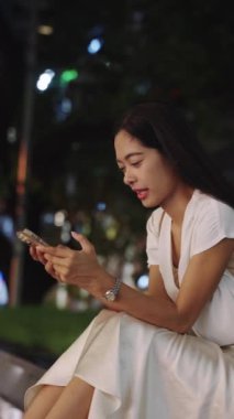 İş kadını gece Bangkok, Tayland 'da dışarıda oturuyor, akıllı telefonunu kullanırken gülümsüyor.