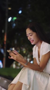 İş kadını geceleri dışarıda oturuyor, Bangkok 'un canlı şehrinde akıllı telefon kullanırken gülümsüyor.