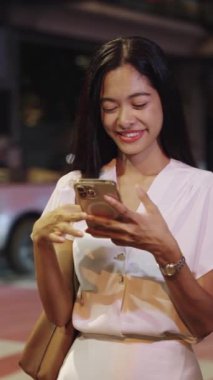 İş kadını gece Bangkok 'ta akıllı telefon kullanırken gülümsüyor, canlı şehir atmosferinin tadını çıkarıyor.