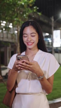 Genç iş kadını geceleri Bangkok sokaklarında telefonuyla gülümsüyor.