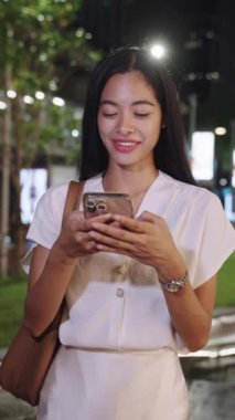 Genç iş kadını gece Bangkok 'ta kaldırımda duruyor, telefonuna mesaj atarken gülümsüyor.