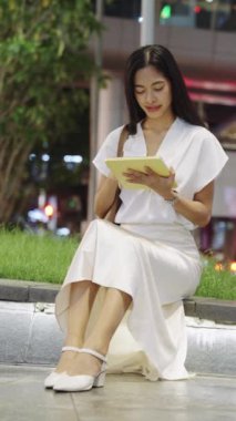 Genç iş kadını geceleri Bangkok 'ta alçak bir duvarda oturuyor, bir tablet üzerinde çalışıyor ve mola sırasında esniyor.